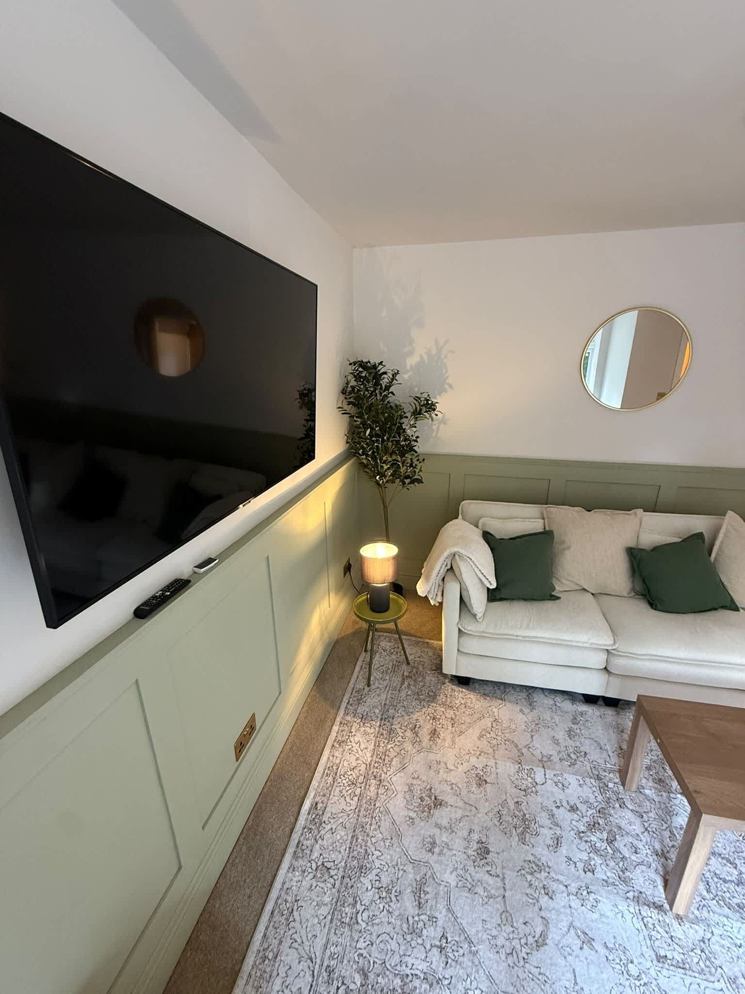 Una sala de estar con una televisión de pantalla plana montada en la pared, un sofá con cojines, una mesita, una lámpara de pie y una planta en maceta.