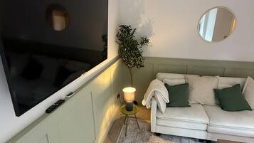 Una sala de estar con una televisión de pantalla plana montada en la pared, un sofá con cojines, una mesita, una lámpara de pie y una planta en maceta.