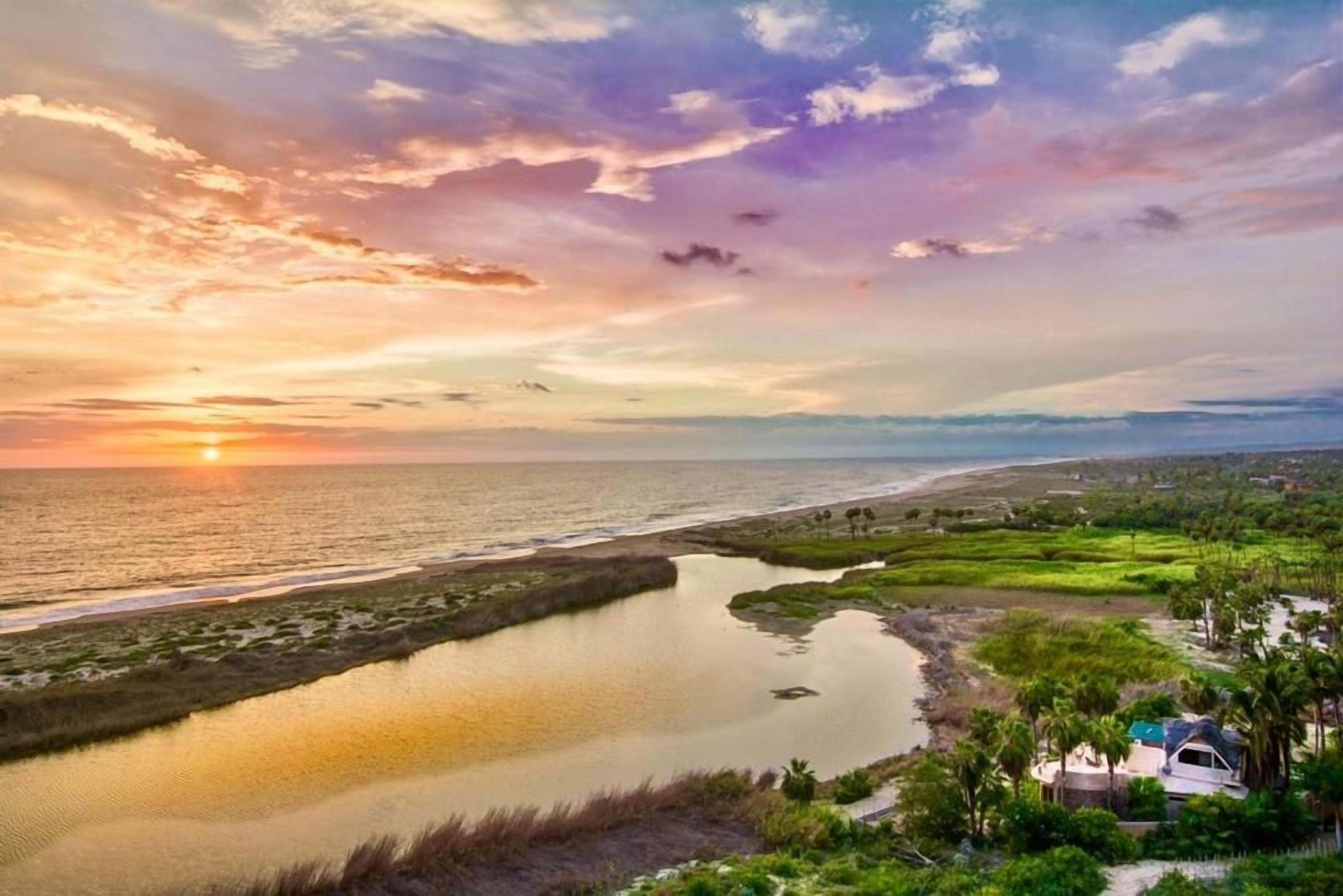 A coastal landscape with a sunset, a body of water, and a small building with a roof.