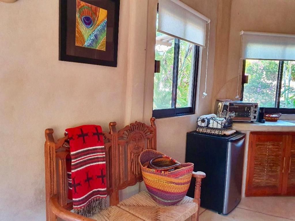 Une pièce comprenant une chaise en bois, un panier en osier, une couverture rouge, un tableau encadré, une fenêtre et un coin cuisine.