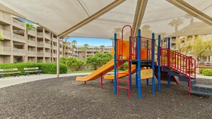 A playground with a slide and climbing structures, surrounded by a covered area and a building with balconies in the background.