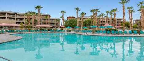 A swimming pool with lounge chairs and umbrellas, surrounded by palm trees and multi-story buildings.