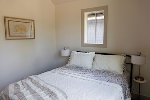 A bedroom with a bed, a window, a wall-mounted lamp, and a framed picture.