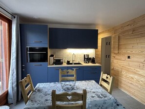 A compact kitchen with blue cabinets, a dining table with chairs, and a wooden accent wall. - 350m from ski slopes, 3rd floor, balcony, parking, tv, ski locker, 36m², Valloire (Valloire)