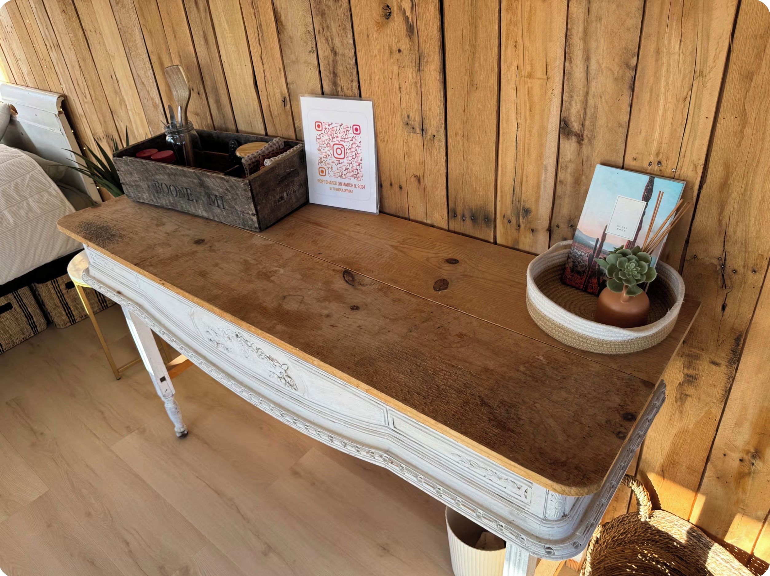 Ein rustikaler Holztisch mit weiß gestrichenem Untergestell, ein kleiner Blumentopf und ein gerahmtes Bild.