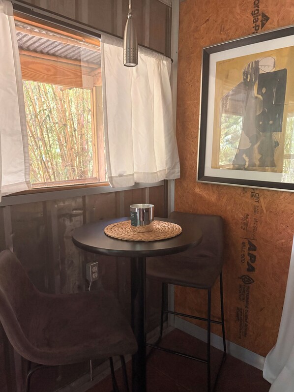 A small room with a table, two chairs, a hanging light fixture, a window with white curtains, and a framed artwork on the wall.