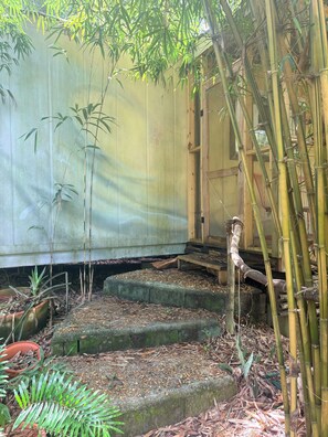 A traditional bamboo structure with a wooden staircase leading up to a door, surrounded by bamboo plants and a small garden area.