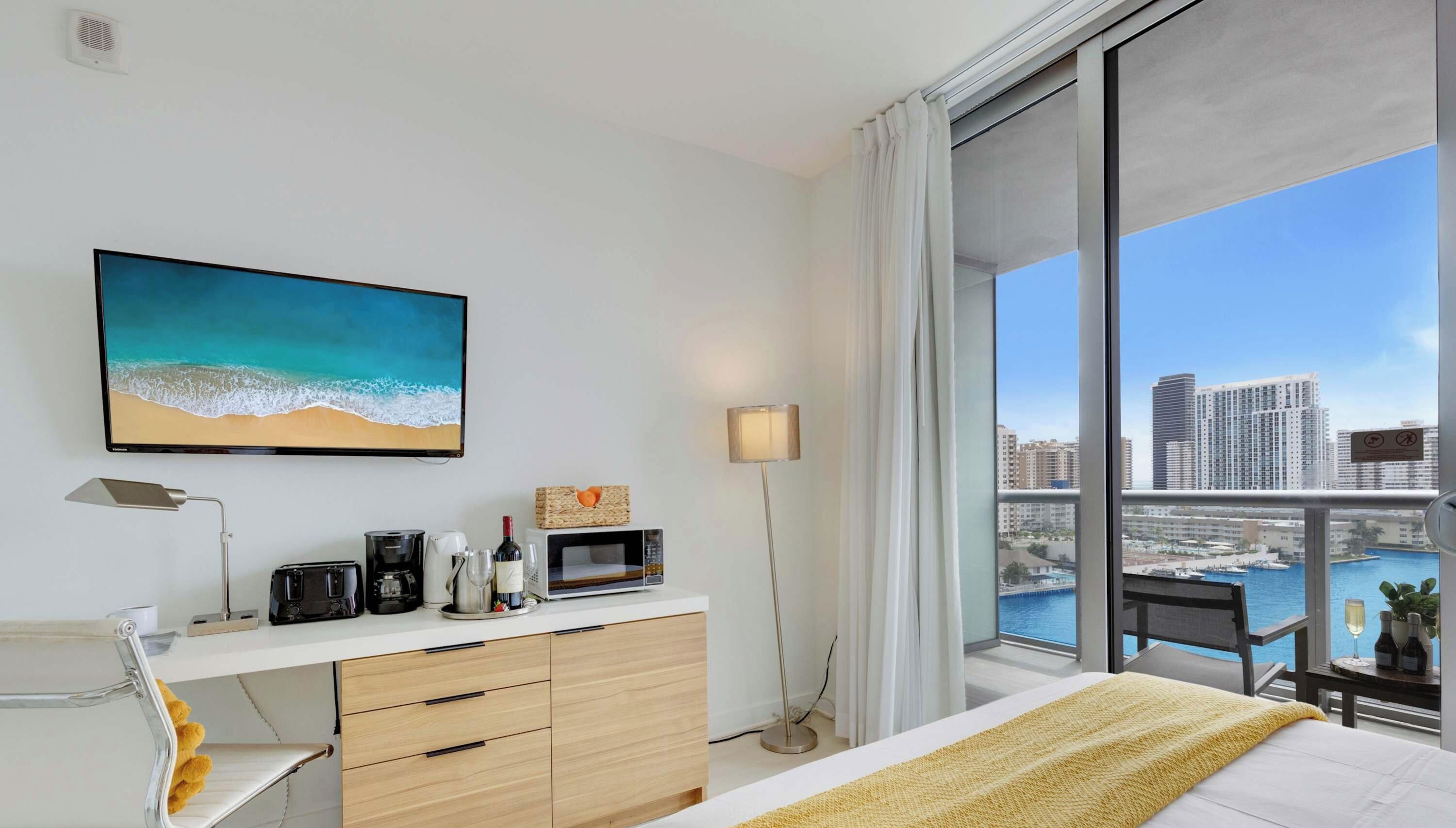 Habitación de hotel con un ventanal grande, cama, escritorio con lámpara, cafetera, microondas y un cuadro de playa.