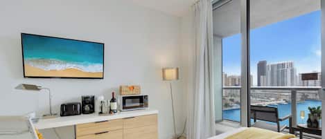 Une chambre d’hôtel avec une grande fenêtre, un lit, un bureau avec une lampe, une machine à café, un four à micro-ondes et un tableau représentant une plage.