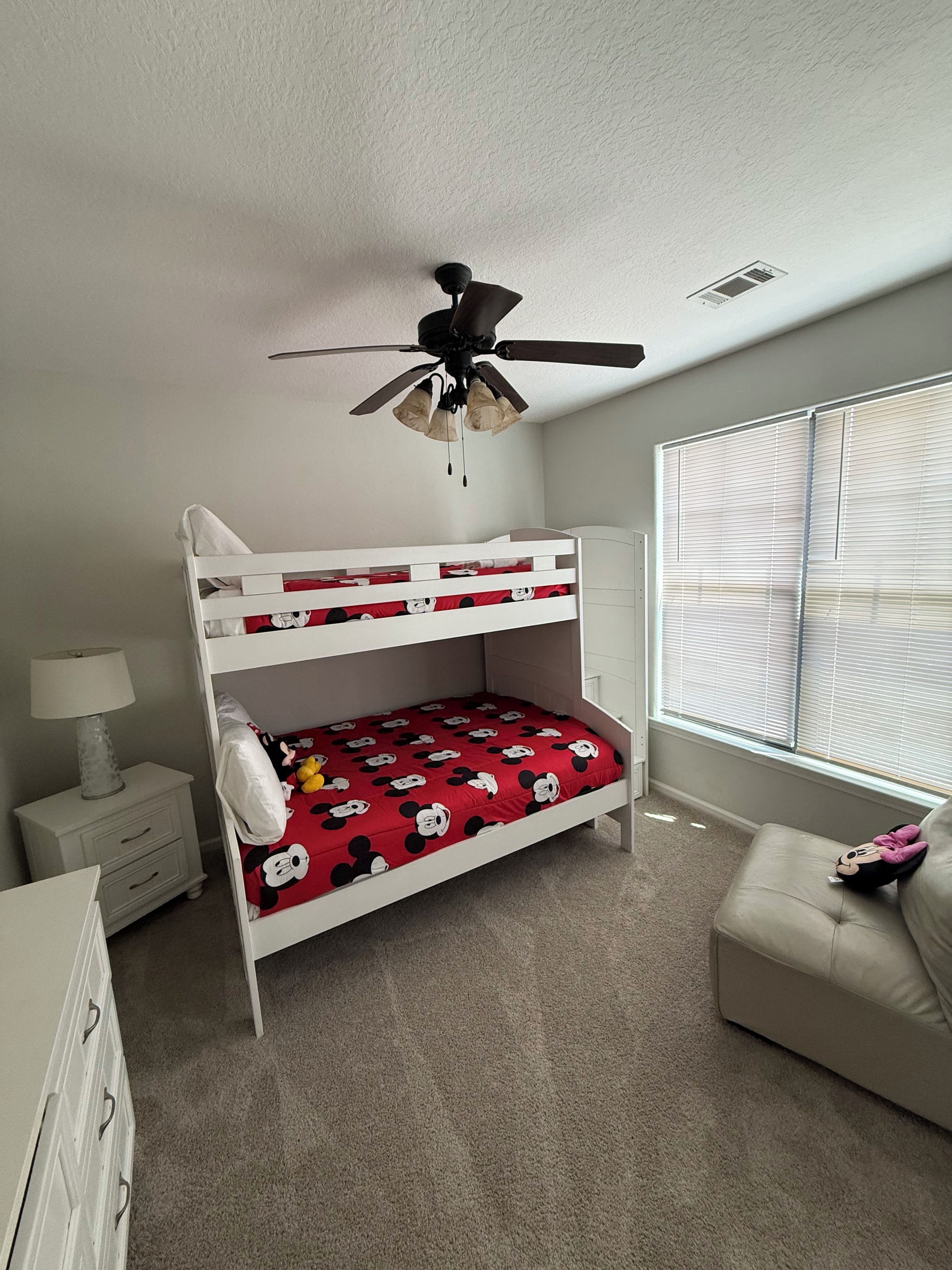 A bunk bed with Mickey Mouse themed bedding, a ceiling fan, a bedside lamp, a window with blinds, and a beige sofa.
