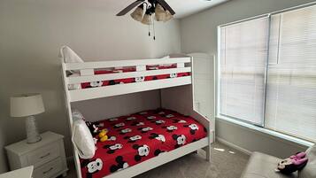 Een stapelbed met beddengoed met Mickey Mouse thema, een plafondventilator, een bedlampje, een raam met jaloezieën en een beige bank.
