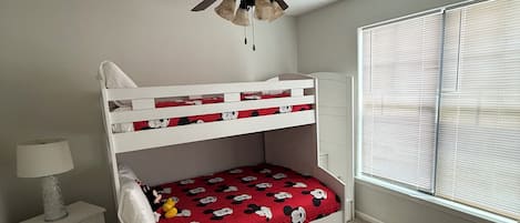 A bunk bed with Mickey Mouse themed bedding, a ceiling fan, a bedside lamp, a window with blinds, and a beige sofa.