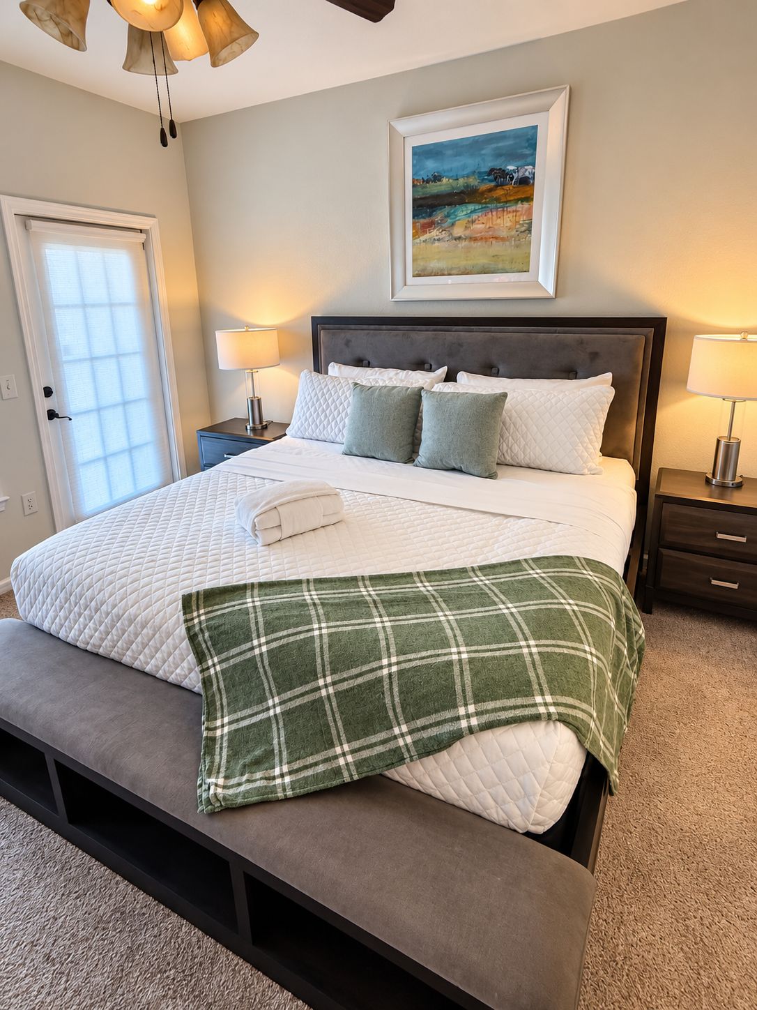 A neatly made bed with a quilted white cover, two pillows, and a green plaid throw blanket. There is a framed painting above the bed, and two bedside lamps on either side.