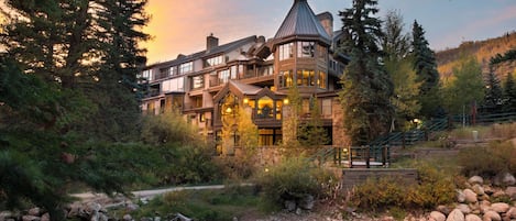 A large, multi-story lodge with a prominent tower, surrounded by dense forest and a flowing stream.