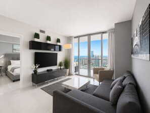 A modern living room with a flat-screen TV, a sofa, a coffee table, and a balcony with a city view. - ★★★★★ Sky-High Oceanview Retreat / Beachwalk Penthouse (Hallandale Beach)