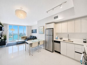 A modern kitchen with stainless steel appliances, a dining area with a table and chairs, and a view of a cityscape through large windows. - ★★★★★ Sky-High Oceanview Retreat / Beachwalk Penthouse (Hallandale Beach)