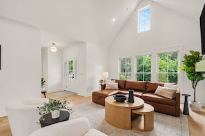 A spacious living room with a brown leather sofa, a round wooden coffee table, and a white armchair. There are large windows providing a view of greenery outside. - Golden Dome Retreat Upper Level Near Notre Dame (South Bend)