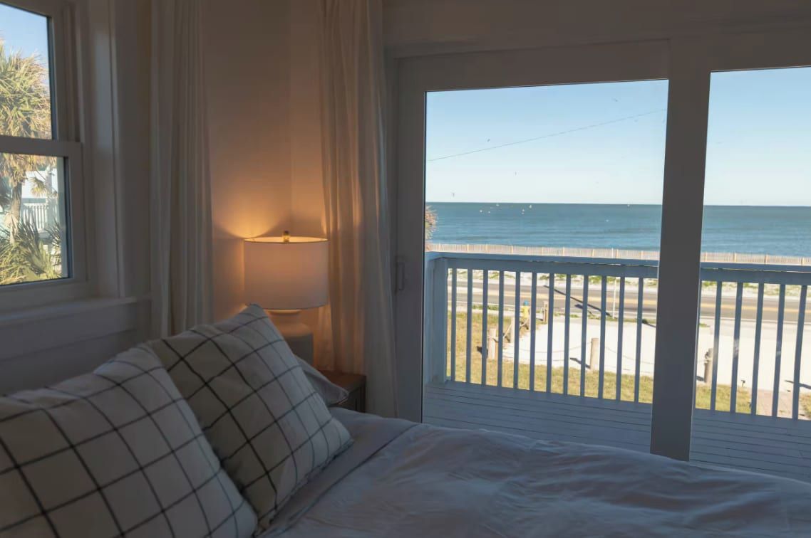 Un dormitorio con cama, mesita de noche, lámpara y vista a la playa desde un balcón.
