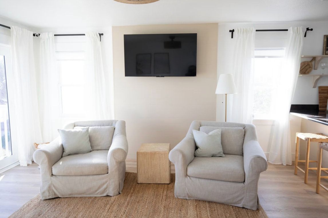 Una sala de estar con dos sillones, una pequeña mesa auxiliar de madera y un televisor de pantalla plana montado en la pared.