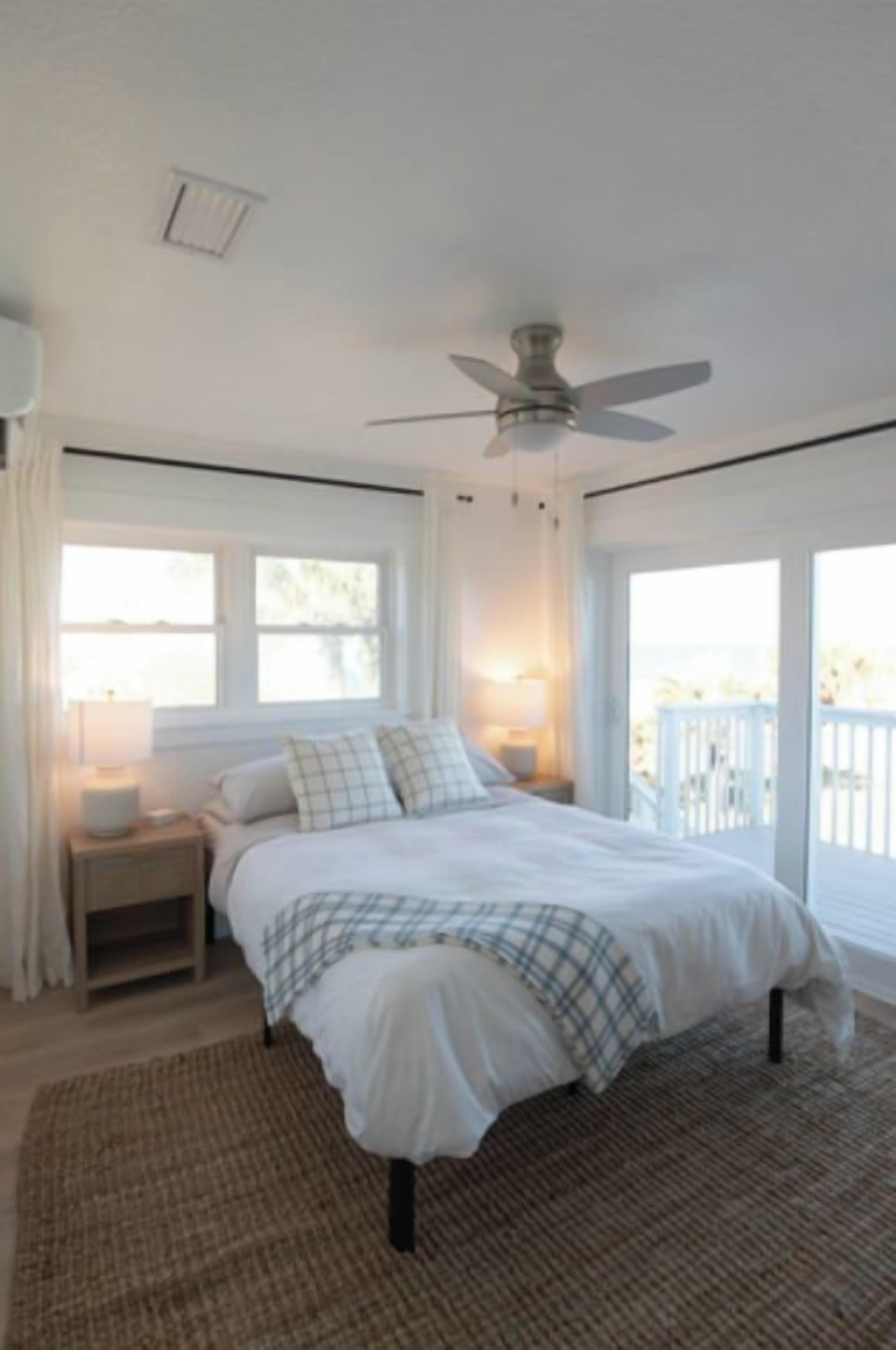 A bedroom with a bed, bedside lamps, a ceiling fan, and a view of the outdoors.