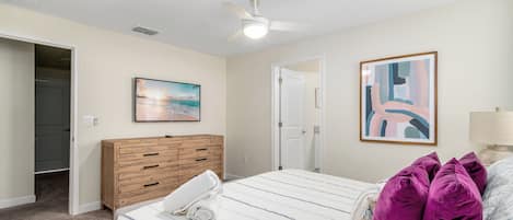 A bedroom with a bed, a wooden dresser, a ceiling fan, a painting, and a door leading to another room.