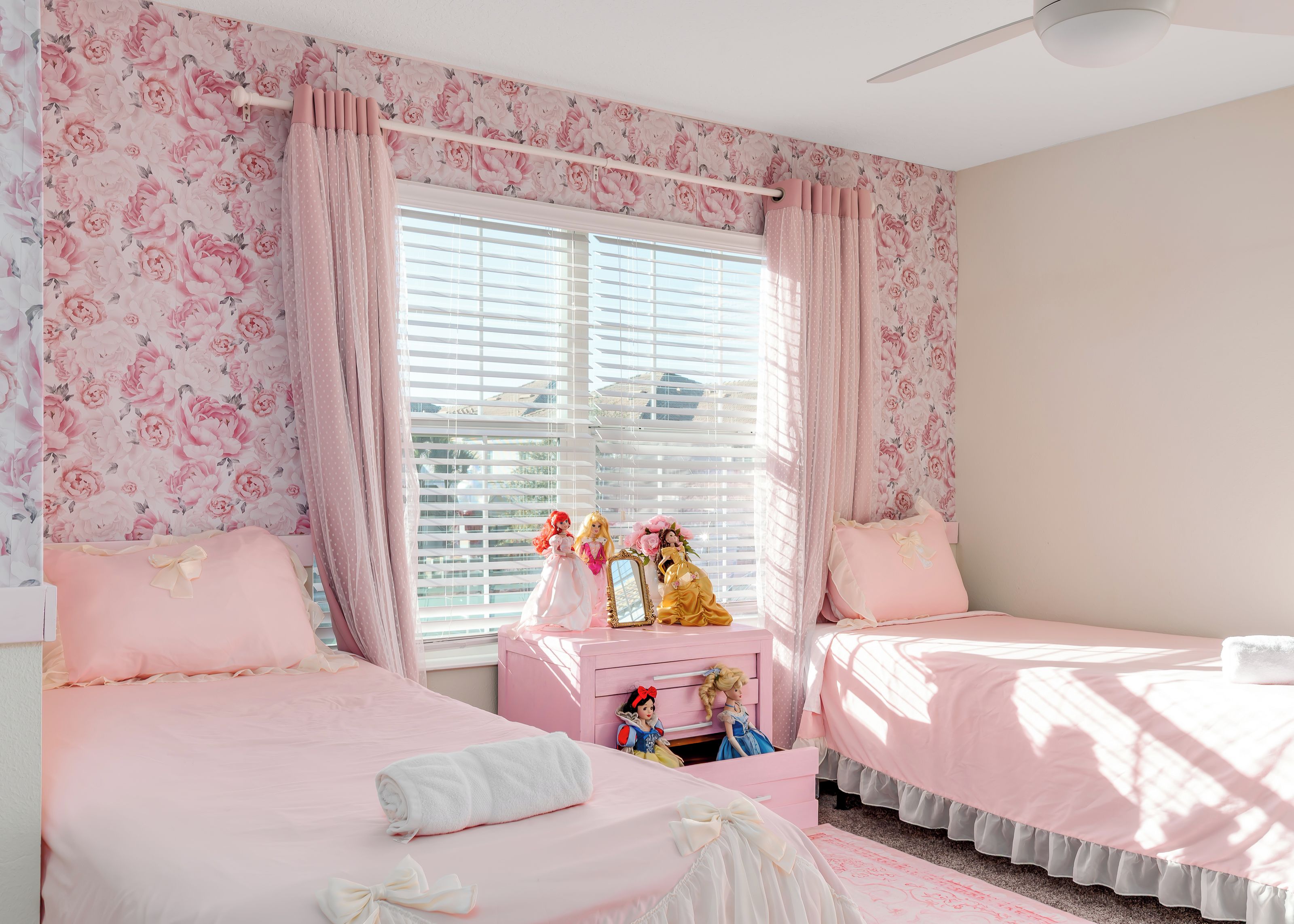 A bedroom with two beds, a vanity with dolls, and floral wallpaper.