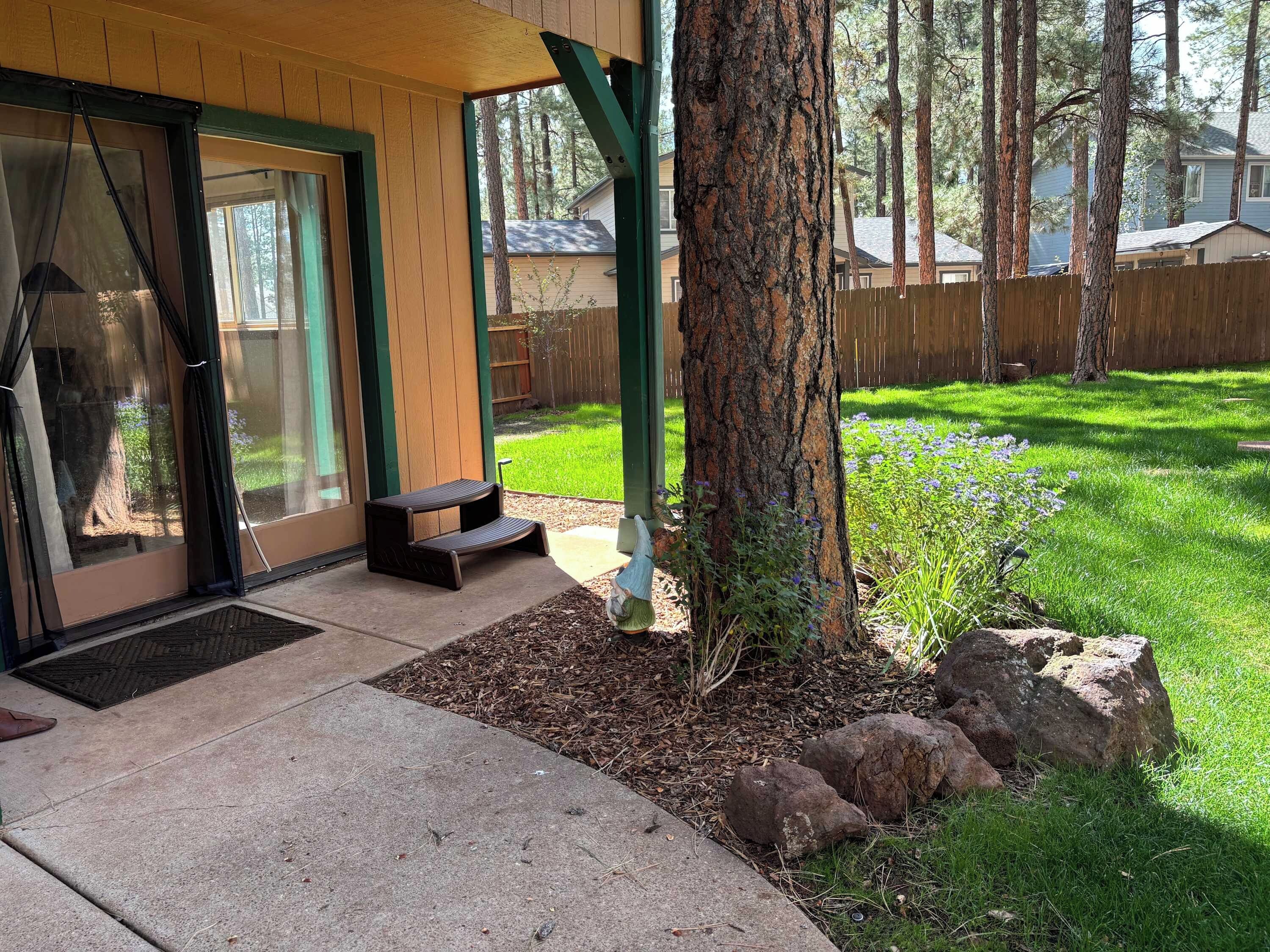 Ein überdachter Patio-Bereich mit einer Bank, einem Baum und einem Rasenstück im Hintergrund, umgeben von einem Holzzaun.