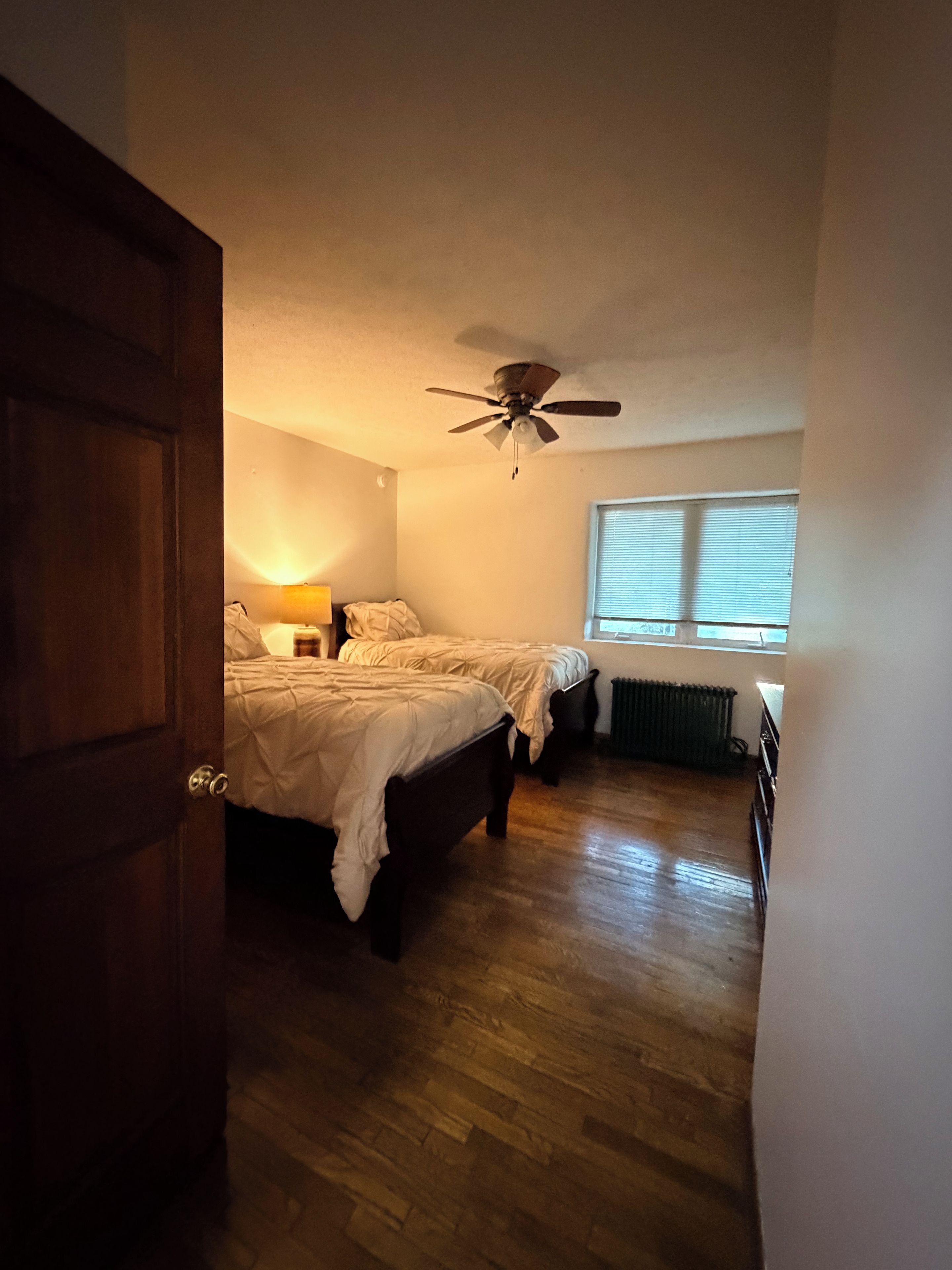 A bedroom with two beds, a ceiling fan, a window with blinds, and a radiator.