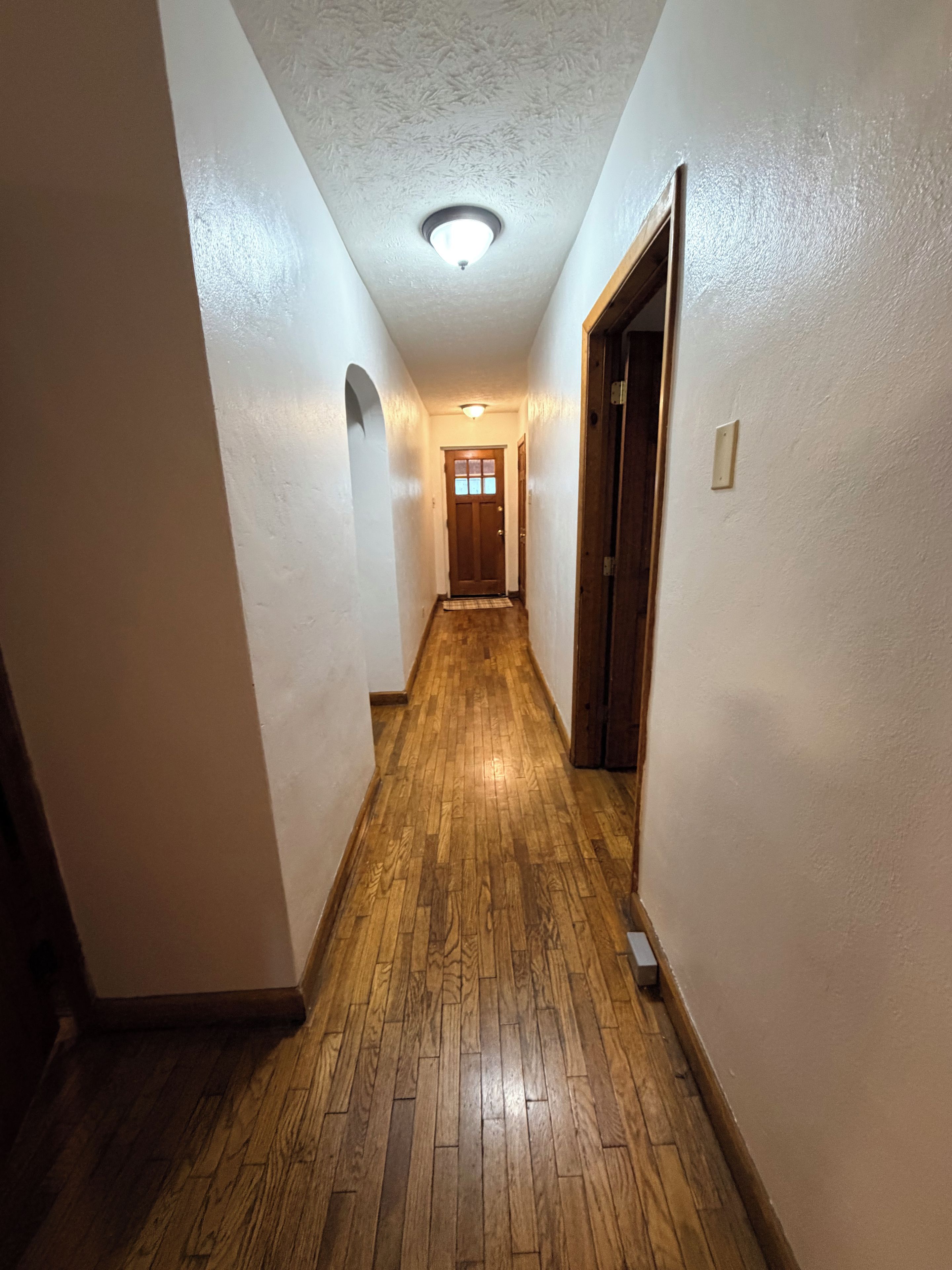 A narrow hallway with wooden flooring and white walls, leading to a door at the end.
