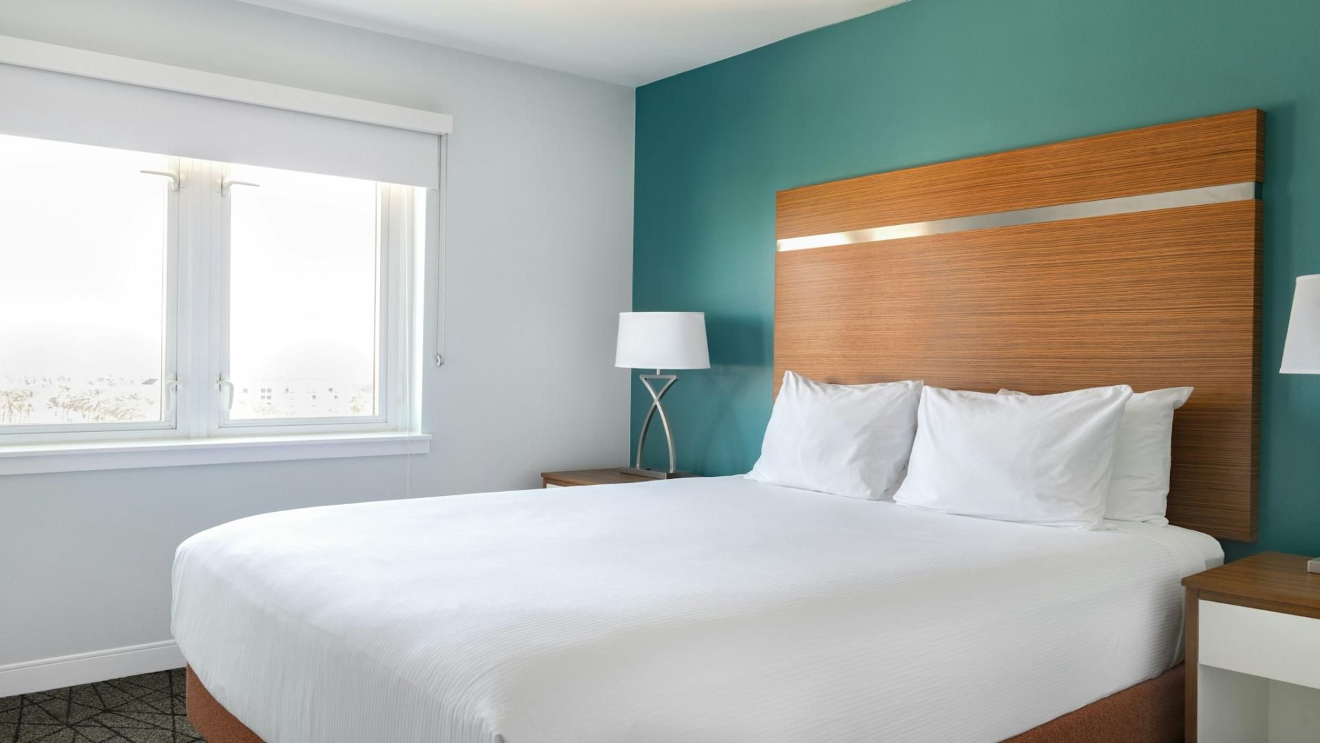 Habitación de hotel con una cama grande, una mesita de noche con lámpara, una ventana con persianas y una pared de acento color verde azulado.