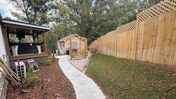 Eine Holzhütte mit Veranda und kleinem Schuppen, umgeben von einem Holzzaun und einem Betonweg.