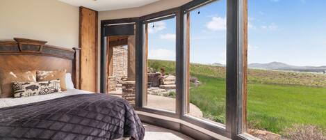 A bedroom with a large bed, a stone fireplace, and a view of a green landscape through large windows.