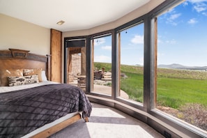 A bedroom with a large bed, a stone fireplace, and a view of a green landscape through large windows.