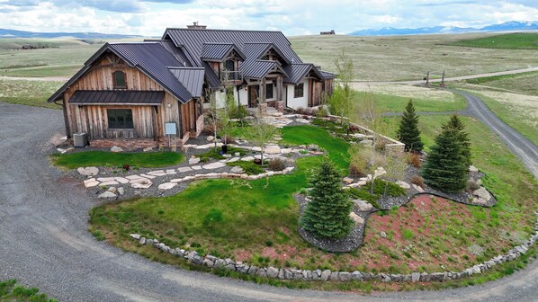 A large wooden house with a metal roof, surrounded by a landscaped garden with a stone pathway, trees, and a view of the countryside.