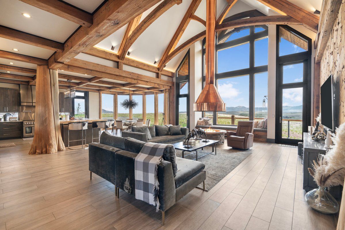 A spacious living area with a large couch, a coffee table, and a fireplace. There is a kitchen area with modern appliances and a large window offering a view of the outdoors.