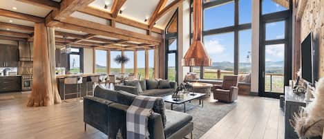 A spacious living area with a large couch, a coffee table, and a fireplace. There is a kitchen area with modern appliances and a large window offering a view of the outdoors.