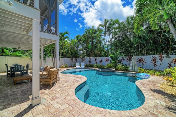 Ein Schwimmbecken mit gebogener Form, umgeben von einer Terrasse mit Gartenmöbeln und einem überdachten Sitzbereich.