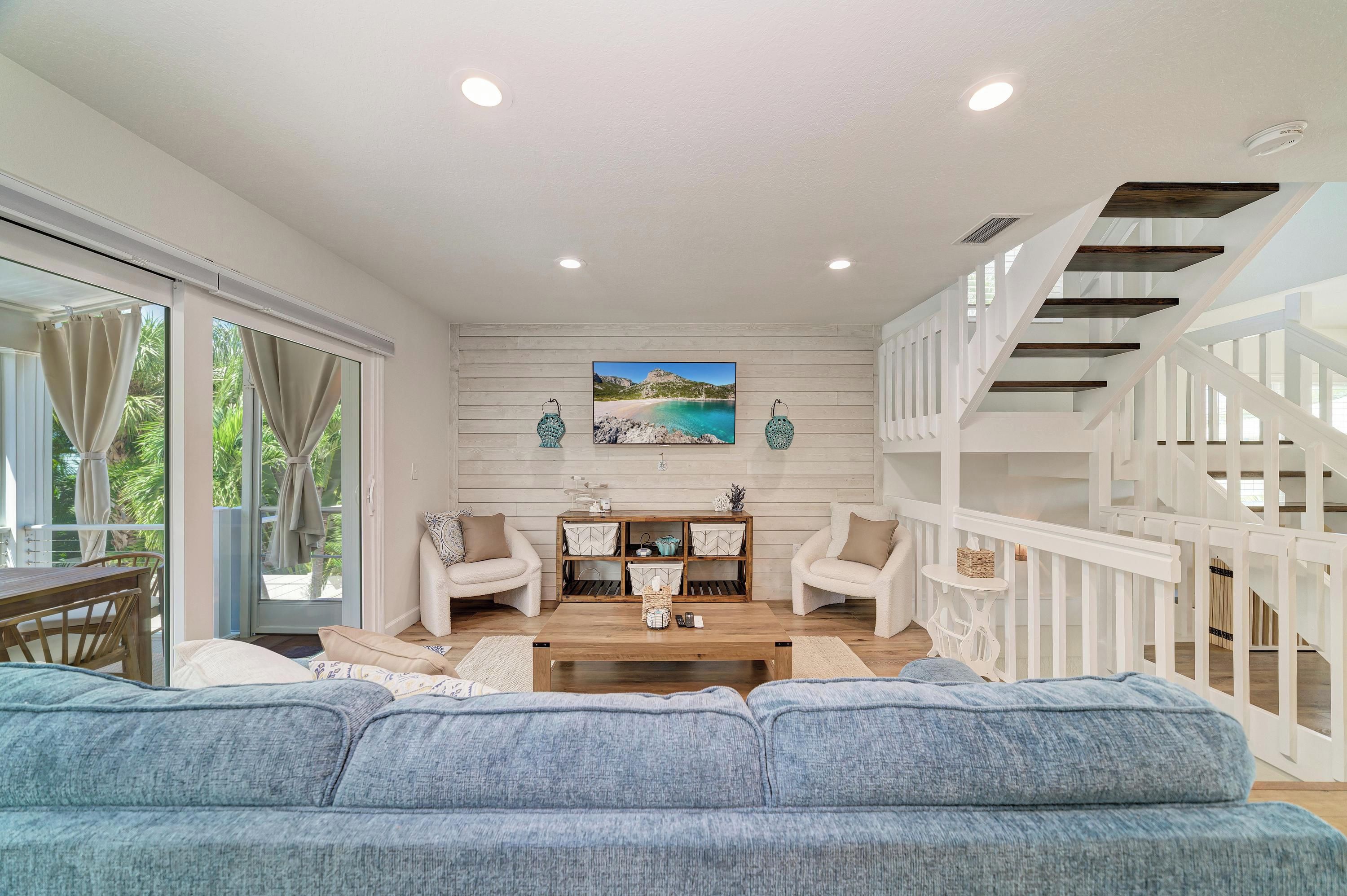 A spacious living room with a large sofa, two armchairs, a coffee table, and a painting of a beach scene. A staircase with white railings is visible to the right.