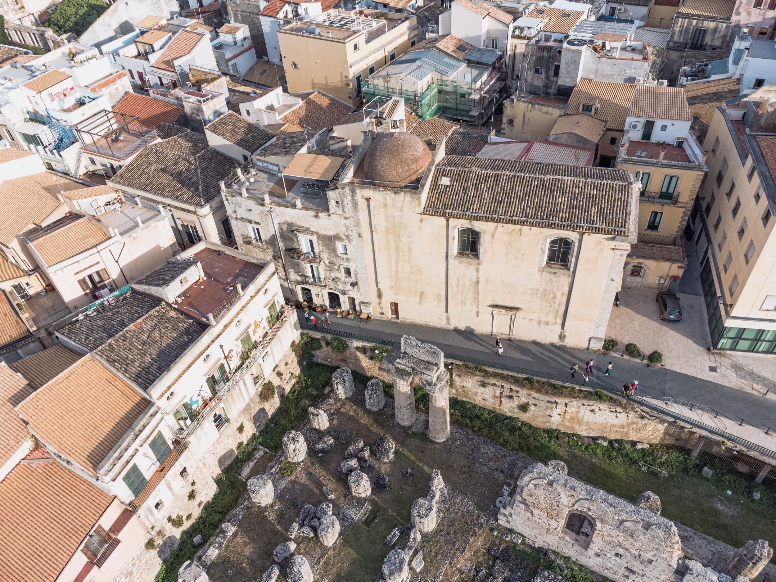 An aerial view of a historic city center with ancient ruins, a prominent church, and tightly packed buildings.