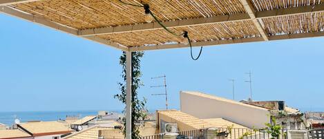 A rooftop terrace with a bamboo roof, outdoor furniture, and a view of the sea.