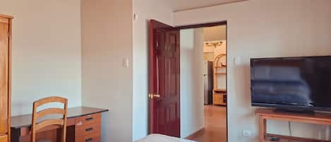 Un dormitorio con cama, escritorio, silla, televisor y puerta que da a otra habitación.