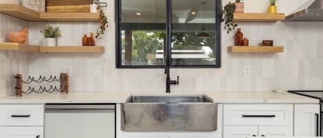 A modern kitchen with white cabinetry, a stainless steel sink, wooden shelves, and a black-framed window.
