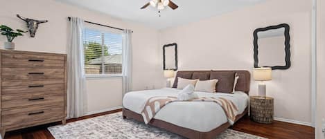 Une chambre à coucher avec un lit, des lampes de chevet, un ventilateur de plafond, une commode et une fenêtre avec des rideaux.