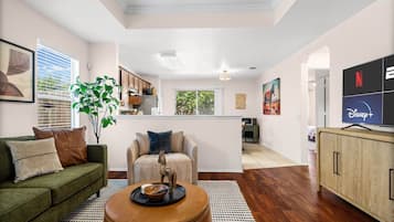 Un salon moderne avec un canapé, une table basse et un téléviseur. On aperçoit une cuisine à l’arrière-plan, avec des placards et une fenêtre.
