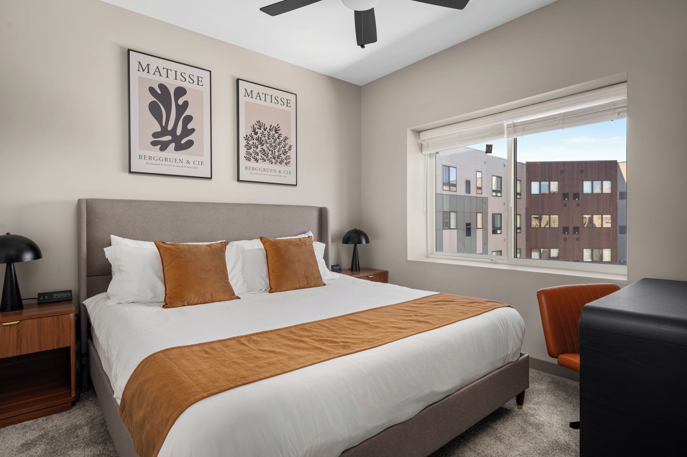 Ein Hotelzimmer mit einem großen Bett, zwei Nachttischen, einem Schreibtisch, einem Sessel, einem Fenster mit Blick auf Gebäude und zwei gerahmten Kunstwerken an der Wand.
