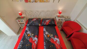 Ein Bett mit Gitarrenmuster auf der Bettdecke, zwei rote Lampen, ein roter Sessel und eine Wand mit Musikinstrumenten.