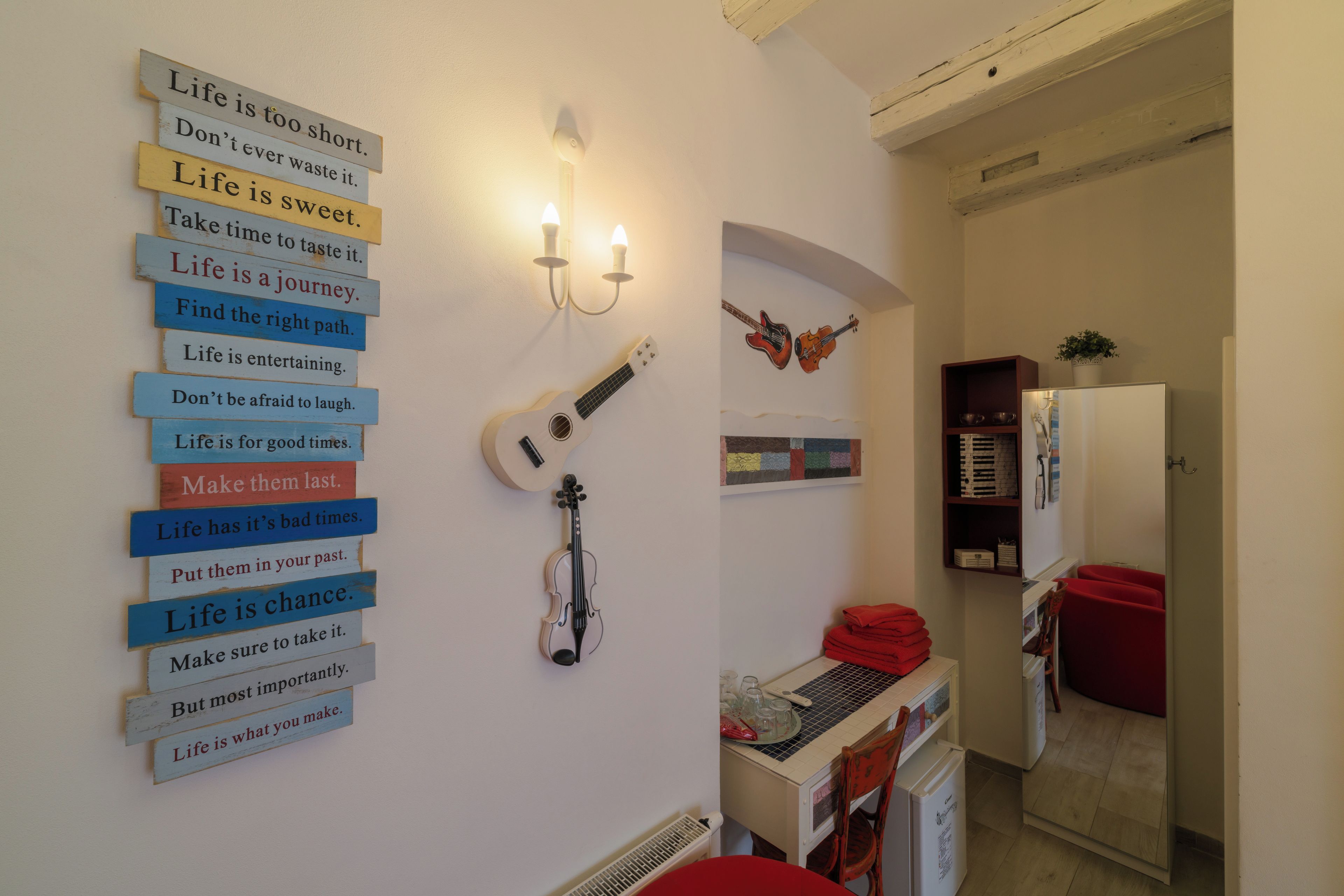 Une pièce avec un luminaire fixé au mur, une guitare et un violon. On y trouve une chaise rouge et une table recouverte d’une nappe rouge.