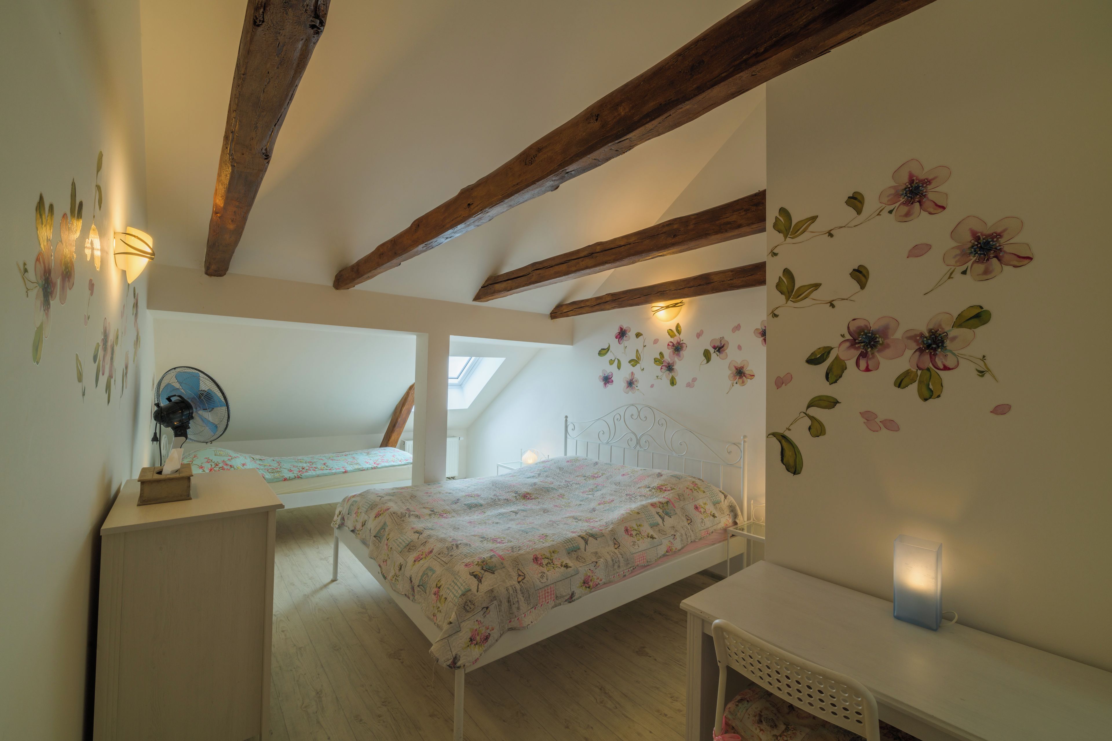 Un dormitorio con techo inclinado, vigas de madera, cama con ropa de cama floral, escritorio con ventilador y pared con adhesivos florales.