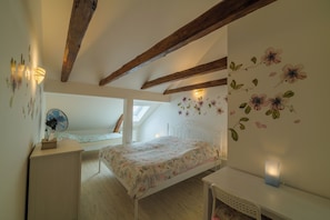 Un dormitorio con techo inclinado, vigas de madera, cama con ropa de cama floral, escritorio con ventilador y pared con adhesivos florales.