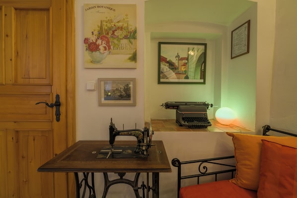 Una habitación con puerta de madera, una máquina de coser sobre una mesa, una cama con un almoháron naranja y varios cuadros en las paredes.